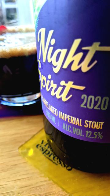 Close-up of Kereru Night Spirit 2020 - barrel aged imperial stout. Purple label, wooden table, yellow coaster next to a glass of black-coloured beer (stout).