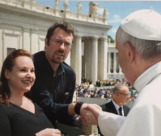 Auf dem Bild ist Papst Franziskus zu sehen, wie er Alexander die Hand schüttelt. Neben Alexander steht Alexa, die lächelnd zuschaut. Im Hintergrund erkennt man den Petersplatz im Vatikan mit Säulen und vielen Menschen.