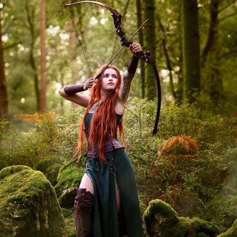 Redhead woman in forest, in a green dress aiming at something above with bow and arrow.
