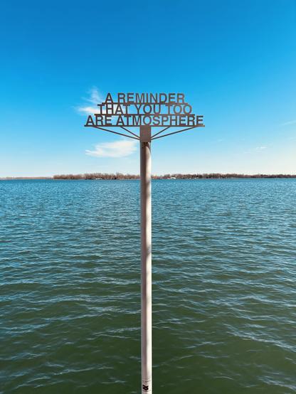 A metal sign on a pole over calm water, displaying the message: "A REMINDER THAT YOU TOO ARE ATMOSPHERE." The background features a clear blue sky with some clouds.