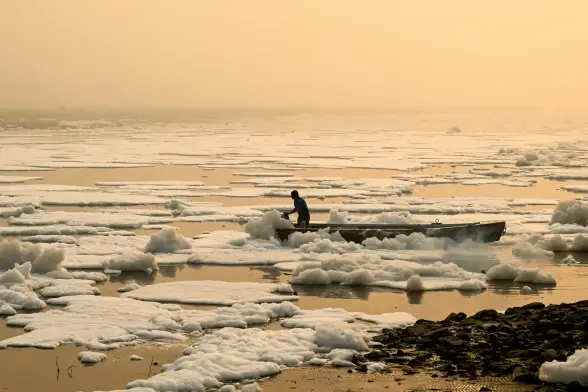 A man in a boat in a haze in a river covered with toxic white foam as far as the eye can see.