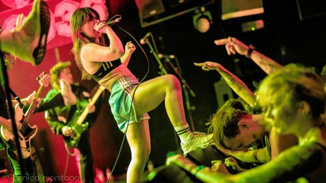 A female singer performs energetically on stage, holding a microphone and singing, while wearing a crop top and shorts. Behind her, band members play instruments. The crowd is engaged, with hands raised and some individuals leaning forward, expressing excitement.