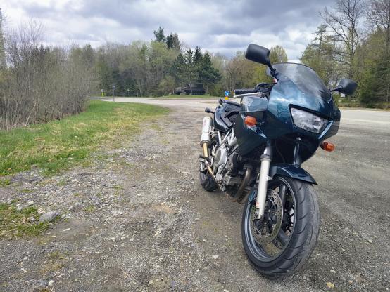 Ein grünes Motorrad steht auf dem Seitenstreifen neben einer Straße geparkt. Im Hintergrund ist ein Panzer auf einem Kreisverkehr zu sehen.