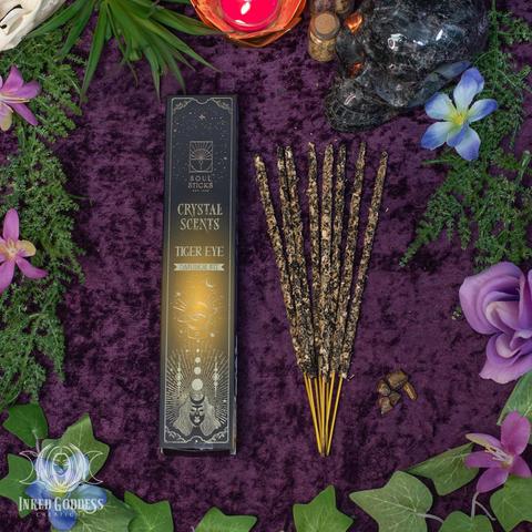 The contents of a box of the "Crystal Scents: Tiger Eye" incense product are arranged on a velvet surface. The long rectangular box is on the left with a fanned array of incense sticks on the right. In the lower right is a small bunch of tumbled Tiger's Eye gemstone chips, included in the box. Presented by Inked Goddess Creations.
