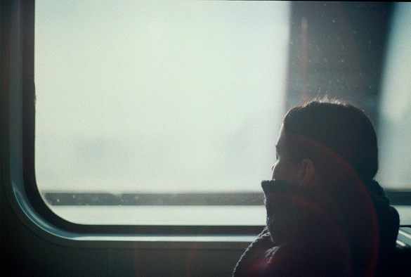 The image depicts a child sitting by a window, likely on a train or bus. The child is facing away from the camera, looking out the window. The window is large and rectangular, with a rounded top, and the view outside is hazy and indistinct, suggesting it might be foggy or the child is looking through a layer of mist. The child is wearing a dark-colored sweater with a high collar, which is turned up, possibly to keep warm. The lighting in the image is soft and diffused, with a lens flare effect visible on the right side, indicating sunlight coming through the window. The overall atmosphere is calm and contemplative, with the child appearing to be in a moment of quiet observation.