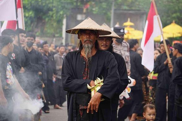 Large group of people in dark clothing, many wearing conical hats, participating in a ceremony, with flags and smoke.