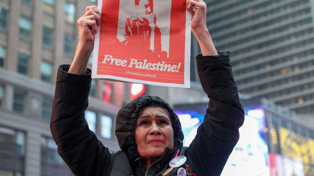 Foto van vrouw die een krant omhoog houdt, voorpagina is in rode opdruk een foto van Mahmoud Khalil, met de tekts "Free Palestine" en daaronder de hashtag #FreeMahmoudKhalil