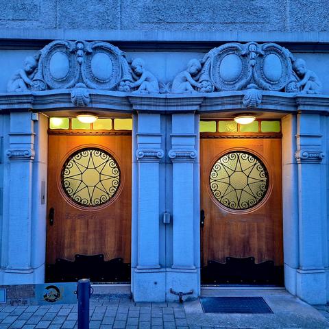 A picture shows twin wooden doors framed by stone columns and topped with stone shields and cornucopias. Four sculpted children reach for fruit spilling from the cornucopias. Circular windows with star-shaped grilles are set into the doors.