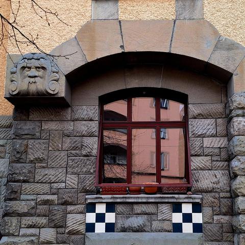 Close-up view of a window on a historic building. The window, located on the ground floor, is set into a roughly hewn stone wall. It is sheltered by the upper part of the building, which slightly overhangs. In the upper left corner, there is a sculpted grotesque figure. Below the window, there are two tiles featuring black and white checkerboard patterns, which appear slightly out of place.