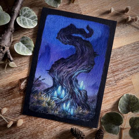 A photo of a portrait-format painting on black paper, placed on a wooden table and surrounded by branches, dried flowers, and leaves for decoration. The painting features a gnarled, twisting tree without branches, standing in a field of dry yellowish grass. Luminescent pale blue mushrooms grow at the tree‘s base, and indistinct mountain-like shapes are visible in the purple/blue, misty background.