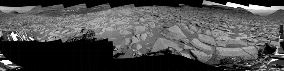 A black and white mosaic capturing a Martian landscape. In detail, NASA's Mars rover Curiosity took 19 images in Gale Crater using its mast-mounted Right Navigation Camera (Navcam) to create this mosaic. The seam-corrected mosaic provides a 360-degree cylindrical projection panorama of the Martian surface centered at 187 degrees azimuth (measured clockwise from north). Curiosity took the images on April 14, 2025, Sol 4510 of the Mars Science Laboratory mission at drive 816, site number 115. The local mean solar time for the image exposures was 2 PM. Each Navcam image has a 45 degree field of view. 