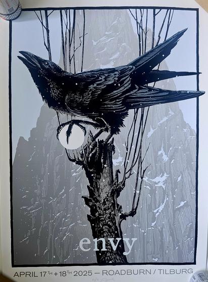 Poster for the band Envy's performance at Roadburn Festival in Tilburg on April 17th and 18th, 2025. The artwork features a dark, intricately detailed raven perched on a gnarled tree branch, holding a glowing white orb in its claw. Thin, leafless branches stretch upward around the bird. The background shows misty, mountainous terrain in shades of gray, creating a moody, atmospheric scene. The word "envy" appears in lowercase at the bottom center in a light serif font.