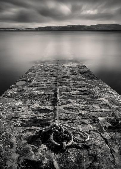 A photograph of a mooring ring and rope extending into the still waters of Loch Ewe, taken from a stone launching ramp near Midtown in the Scottish Highlands. The long exposure smooths the surface of the loch, contrasting with the rough texture of the stone in the foreground. The monochrome tones highlight the simplicity of the scene, emphasising the quiet, coastal atmosphere.