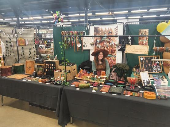 My leathercraft booth in Salon Fantastique in all its glory ! With lots of colored leather goods on black tablecloths, in front of my personalised banner, green backgrounds and myself smiling, wearing my witchhat and outfit for the fair.