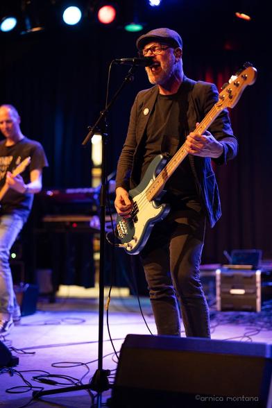 A musician sings into a microphone while playing a blue bass guitar on stage. Another band member is seen in the background, playing guitar. The scene is illuminated by colorful stage lights against a dark backdrop.