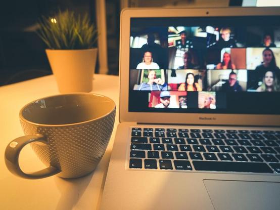 A close-up of a laptop displaying a video conference with multiple people on screen. In the foreground, there is a textured grey coffee mug on a white table, and a small potted plant is visible in the background. Image from unsplash.com