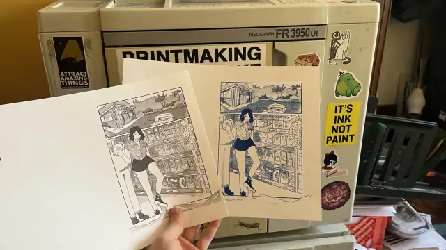 In the foreground, Neve holds a black and white comic page, as well as a blue risograph printed comic page-- on the page, in panel one, a drawing of a trader joe's, in panel two, an annoyed trader joe's worker steps into a mysterious goo as she slaps a bag of chips into a customers chest. Behind the pages, there is an FR Riso covered in stickers. 