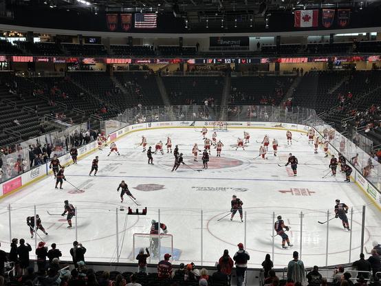 The Coachella Valley Firebirds warm up for their game against the Calgary Wranglers. We see players at various spots on the ice warming up with the Firebirds wearing their dark home jerseys in the foreground and the Calgary Wranglers wearing their road whites in the distance. 