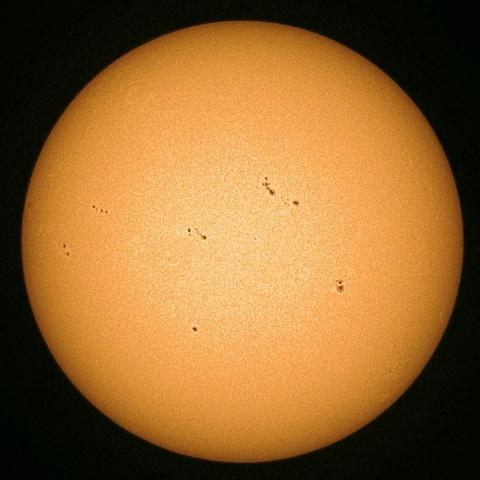 A close-up view of the sun, featuring a bright orange surface with several dark sunspots scattered across it.