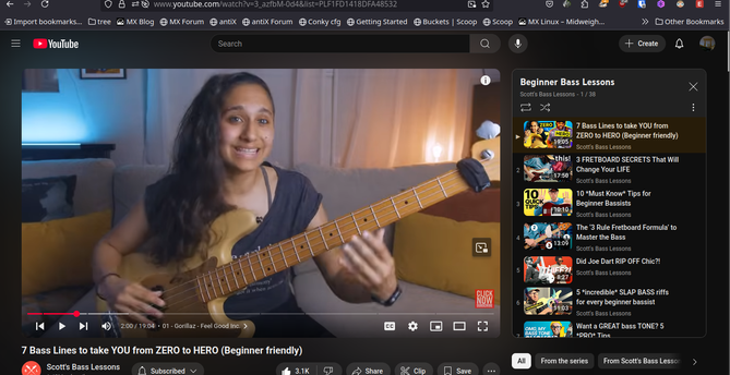 The screencap shows a YouTube video page with a video titled "7 Bass Lines to take YOU from ZERO to HERO (Beginner friendly)" by Scott's Bass Lessons. The video features a woman with long, dark hair, wearing a dark-colored top, sitting on a couch and playing a bass guitar. She is smiling and appears to be in a home setting with a couch, a yellow pillow, and a blue-lit background. The video player interface is visible, including the play/pause button, progress bar, and volume control. The video has 3.1K views and is marked as "Live." On the right side of the screen, there is a sidebar with a list of related videos under the "Beginner Bass Lessons" category, including titles such as "3 FRETBOARD SECRETS That Will Change Your LIFE," "10 'Must Know' Tips for Beginner Bassists," and "The 3 Rule Fretboard Formula to Master the Bass." The YouTube interface includes the search bar, the bell icon for notifications, and the user's bookmarks and subscriptions.

Ovis2-8B