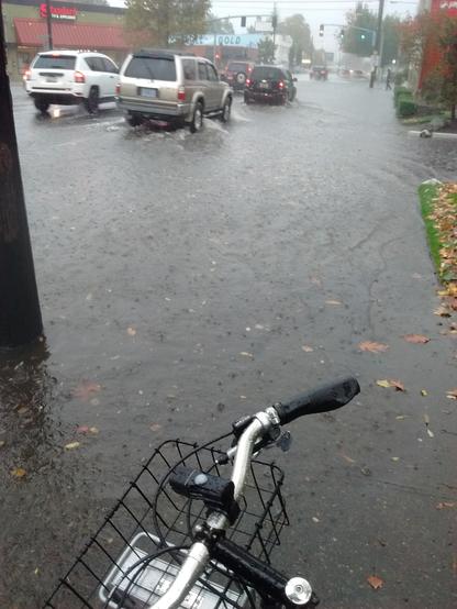 looking past my bike handlebars into a flooded street with traffic driving through the water. It's gray and raining.