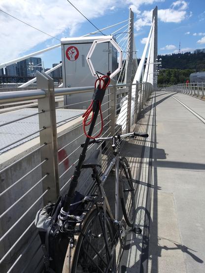 my bike leaning against a wire fence on a pedestrian bridge with a device used for recording electromagnetic waves hastily strapped into a pannier, looking very janky.