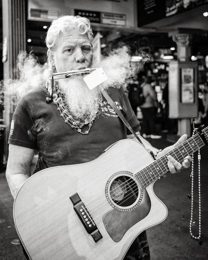 Black and white photo of a man who is blowing vape smoke through his harmonica while holding a guitar.