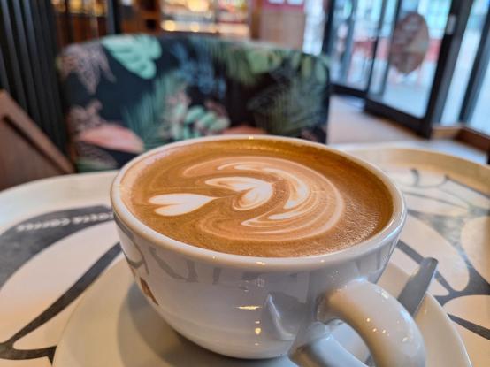 Photo of a white cup with double-heart latte art on top, on a table or tray patterned with white coffee beans on a black background. Blurred background with cafe interior and glass door and large windows to street scene.