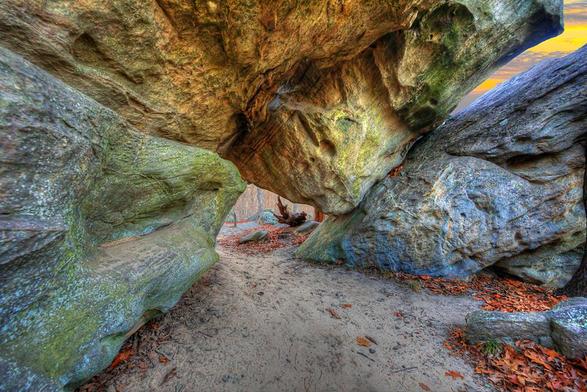 Massive rock formations create a natural archway, framing a sandy path lined with fallen leaves.