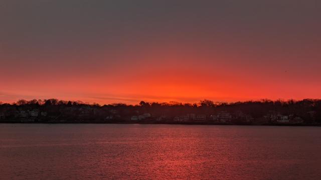 Historical Documentation in the form of a photograph of a rising sun over the ocean bay at the Historic Pioneer Village in Forest River Park, Salem, MA.  The sky is an electric gradient of orange and pink hues, with the sun rising in the distant background. The water is calm and reflects the colors of the sky, creating a mirror-like effect. In the foreground, there are trees silhouetted against the sky. On the right side of the image, there is a small town or village with houses and buildings visible. The overall mood of the photograph is peaceful and serene.