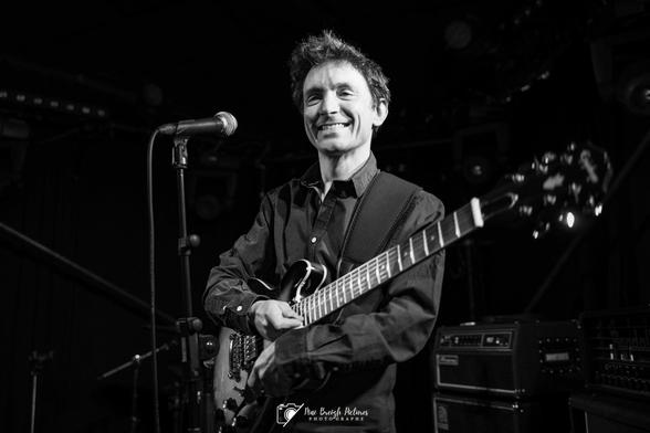Clément Bertine appearing on a black & white photograph, holding a guitar and with a large smile.