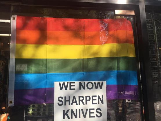 Pride flag hung in store window. In front of it is a sign which reads "we now sharpen knives."
