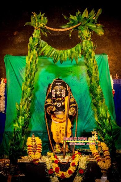 Black stone sculpture of Hindu deity Krishna in yellow pants-like bottoms, gold ornaments, gold shoes, wooden staff in left hand, standing under a crude arch made of green mango leaves. A small gold idol on either side of Krishna, each with yellow flower garland around them. Neon green cloth background.