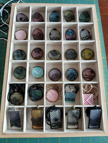 A velvet beige jewelry tray with small bins for items. There are five columns across and six rows down. The upper half is mostly oval and round brooches with Ipswich lace (black or white) on the surface and a glass with a color behind it. 

The bottom two rows are the necklaces with square or rectangular pendants but the same basic thing. Lace applied to the glass surface and a color underneath. 