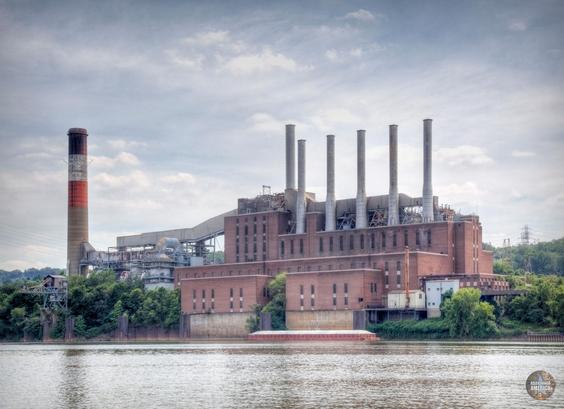 View across a body of water with a massive brick power plant on the opposite shore, crowned by six smokestacks with another larger smokestack off to the side. The plant towers over the trees surrounding it.