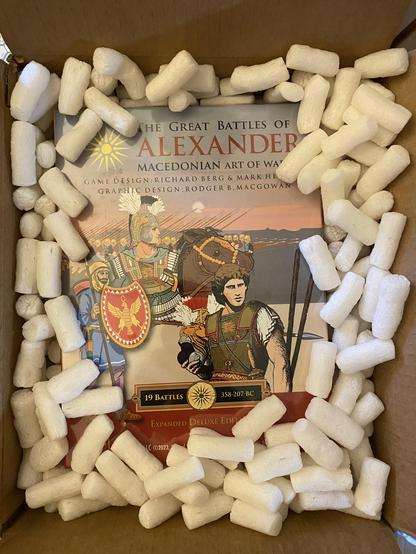 A picture of the board game The Great Battles Of Alexander, the Macedonian Art of War by GMT games. The box is still inside the shipping box and is slightly obscured by packing peanuts. In the center is a drawing of Alexander. 