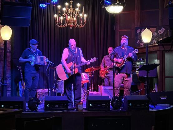 An acoustic band playing on a stage under a big chandelier and moody stage lighting. From left to right there is a button accordion player, an acoustic guitar player, a bass player in the back, and a mandolin player on the right who appears to be singing. Behind the guitar player is a drum kit but the view of the drummer is blocked.