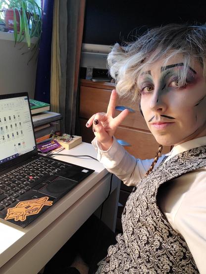 A photo of Austin Tacious in pale pop-art makeup with colourful eyeshadow, making a peace-sign and sitting at his desk where his computer has lots of files open. Something is brewing.