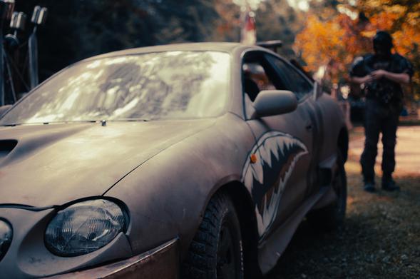A dusty, armored car with a shark-mouth motif painted along the side sits parked on sun-dappled grass. The vehicle is heavily modified for a post-apocalyptic aesthetic, with reinforced bumpers, thick tires, and a matte finish dulled by time. In the background, slightly out of focus, a person in worn tactical gear adjusts something on their chest. Autumn-colored trees cast long shadows, giving the scene a quiet, war-torn beauty.