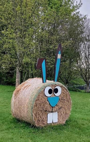 Ein runder Strohballen liegt auf dem Rasen. Ohren, Augen, Nase und Hasenzähne aus Pappe angesteckt, so dass es wie ein Hase aussieht.