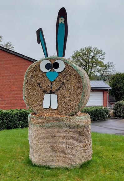 Zwei runde Strohballen aufeinander gestapelt. Ohren, Augen, Nase und Hasenzähne aus Pappe angesteckt, so dass es wie ein Hase aussieht.
