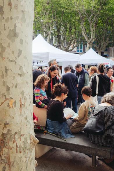 Fotografía de un grupo de adultos y niñes sentados de espaldas en un banco en Plaça Santa Anna en Mataró. Están mirando un libro y sonríen. De fondo, se ven los puestos de la feria de Sant Jordi.
Photograph of a group of adults and children sitting back to back on a bench in Plaça Santa Anna in Mataró. They're looking at a book and smiling. In the background, the stalls of the Sant Jordi fair can be seen.