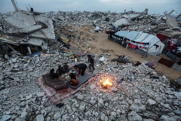 A family has a meal on a carpet by a fire on the ruins of their house, surrounded by rubble & ruins as far as the eye can see.