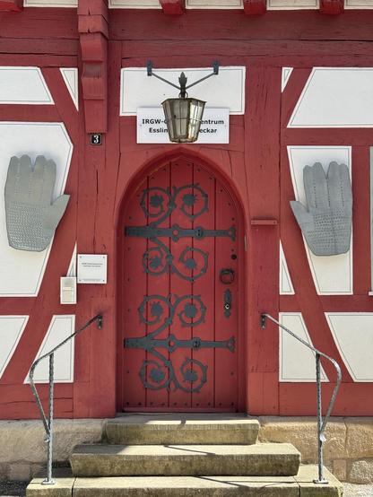 Rote Holztür mit schwarzen Schmiedebeschlägen, darüber eine Laterne. Rechts und links jeweils eine silberne Metallhand. 3 Stufen mit Geländer führen hinauf in die Alte Synagoge in Esslingen
