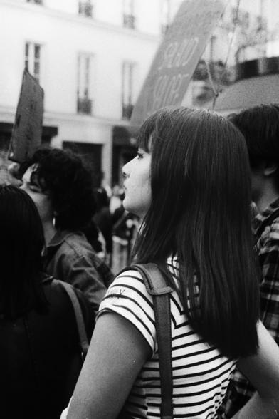 Jeune femme en noir et blanc, au cœur d’une manifestation. De profil, concentrée, elle regarde vers l’avant, l’air calme et attentif. Autour d’elle, la foule s’agite, mais elle reste posée, à l’écoute du monde.
