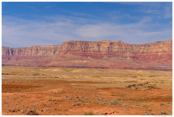 Panoramic view of the Vermillion Cliffs near Page, Arizona, showing layered red and orange rock formations rising above a dry, sparsely vegetated desert landscape under a blue sky with wispy clouds.
