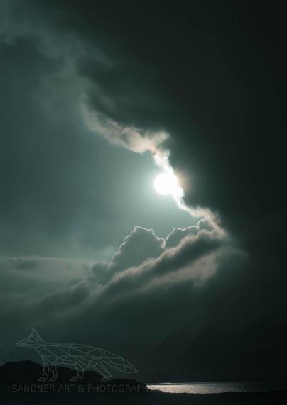 A dramatic photograph of the moon breaking through dark, stormy clouds over Reynisfjara Beach, Iceland. The moonlight casts a silver glow, illuminating the sky and reflecting on the ocean below, creating a striking contrast with the ominous clouds. A watermark reading "Sandner Art & Photography" is visible in the bottom left corner.

