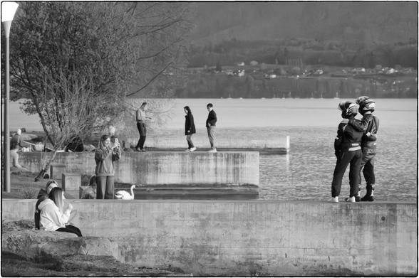 Schwarzweissfotografie verschiedener Leute auf Betonverbauungen an einem Seeufer. Im Zentrum steht ein paar in Motorrad-Vollmontur, das von einer Frau mit dem Smartphone fotografiert wird. 
