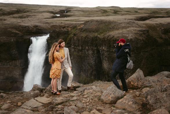 This image captures a behind-the-scenes moment of Chris, an European elopement photographer, during photoshoot in Iceland's dramatic landscape. A couple stands together on rocky terrain beside a stunning white waterfall that cascades down a dark cliff face. The woman wears a bright yellow floral dress with her long hair flowing in the wind, while the man is dressed in a light beige suit with a yellow shirt. To the right, Chris, wearing a red cap and dark coat is capturing their moment, camera raised to his eye. The background features Iceland's distinctive rugged terrain - brown and gray rocky hills with sparse vegetation under an overcast sky. The scene beautifully contrasts the elegance of the couple against the raw, natural power of Iceland's wilderness.