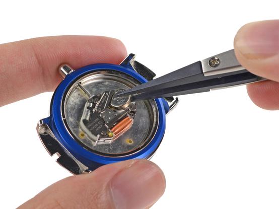 Underside of the Apple analog watch with its bottom cover removed, exposing its internals. This includes a small coin-cell battery, which is being grabbed by tweezers and pulled out of its socket.